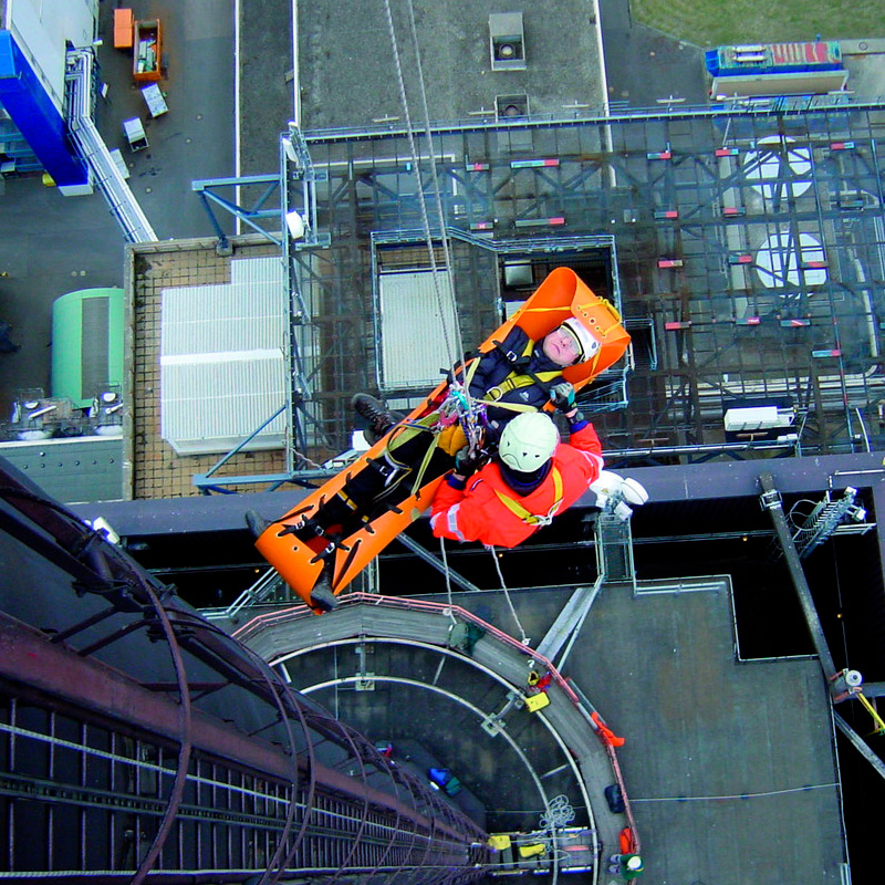 Rescatista bajando víctima por cuerda en ambiente industrial con camilla en posición horizontal.